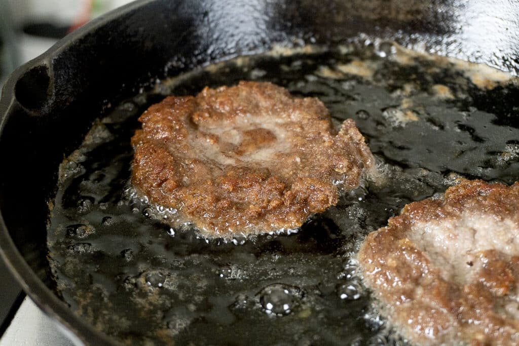 Two slugburger patties are frying in a cast iron skillet with hot oil bubbling around them.