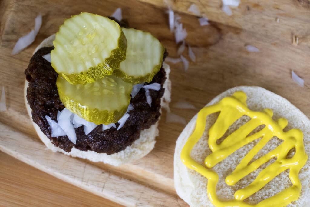 Open slugburger bun with a beef patty, chopped onions, and pickle slices on one half, and yellow mustard on the other, on a wooden surface.