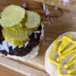 Open hamburger bun with a beef patty, chopped onions, and pickle slices on one half, and yellow mustard on the other, on a wooden surface.