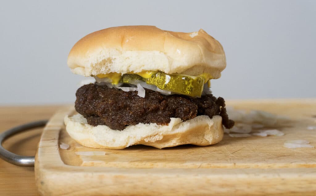 A slugburger with a beef patty, pickles, onions, and mustard on a bun sits on a wooden cutting board.