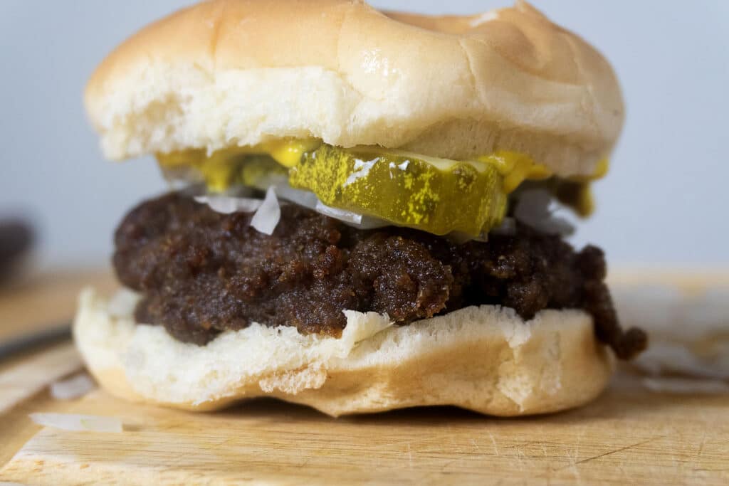 A close-up of a slugburger on a bun with a beef patty, sliced pickles, chopped onions, and yellow mustard, set on a wooden surface.