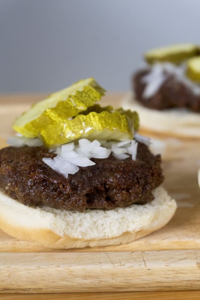 A close-up of a slugburger slider on a bun, topped with chopped onions and sliced pickles, sitting on a wooden surface. Another slider is blurred in the background.