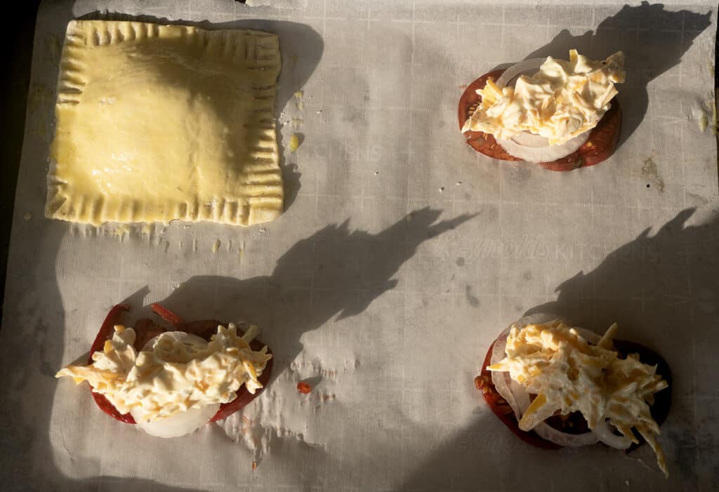Four unbaked pastry items on parchment: one sealed square pastry, and three open stacks with tomato, onion, and shredded cheese, all casting long shadows in bright sunlight.