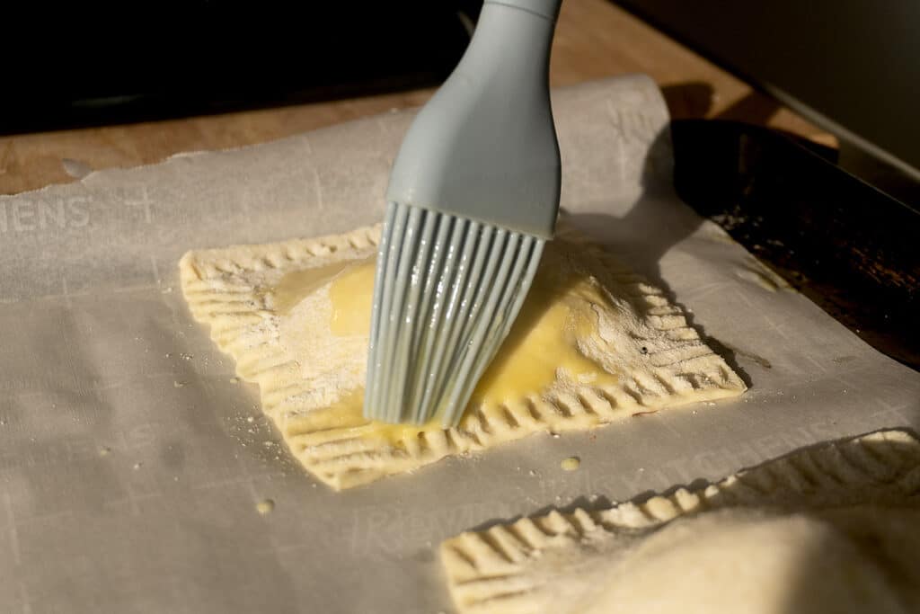 A close-up of a hand brushing egg wash onto a rectangular pastry with a silicone brush on parchment paper, preparing it for baking.