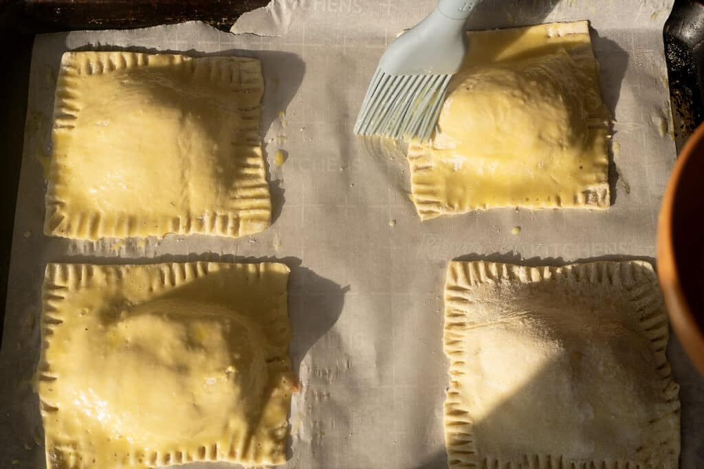 Four uncooked hand pies on parchment paper, with a blue pastry brush applying egg wash to one. Sunlight casts shadows across the scene.