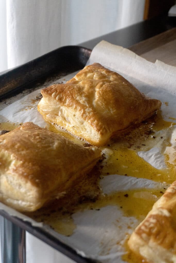 Three golden-brown puff pastry squares rest on parchment paper on a baking sheet, with melted butter or filling pooling around their flaky edges.