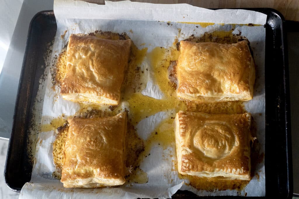 Four golden-brown puff pastry squares on a parchment-lined baking sheet, with some melted butter or filling visible around them.