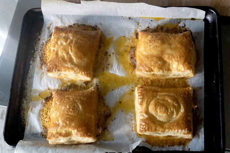 Four golden-brown puff pastry squares on a parchment-lined baking sheet, with some melted butter or filling visible around them.