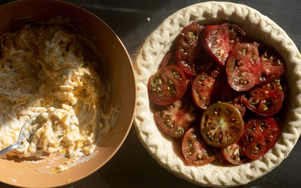 A bowl of cheese mixture with a spoon sits next to an unbaked pie crust filled with sliced tomatoes on a dark surface, lit by sunlight.