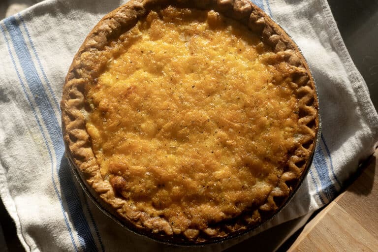 A golden-brown baked pie sits on a white towel with blue stripes, with a fluted crust and a cheesy, bubbly top, lit by sunlight.