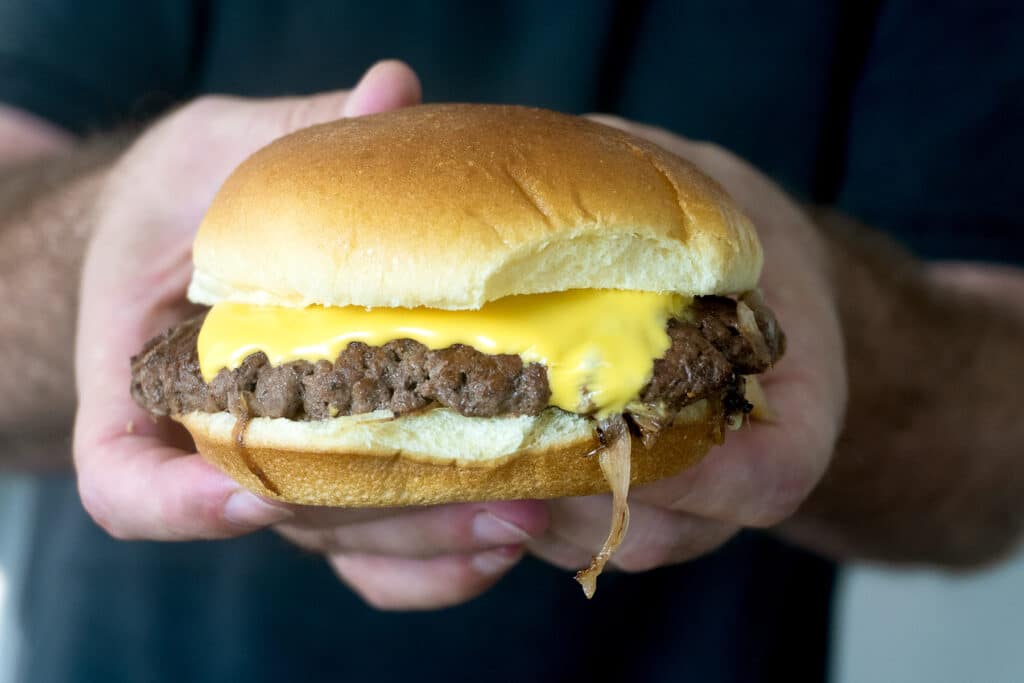 A person holds a cheeseburger with a beef patty, melted cheese, and a soft bun in both hands.