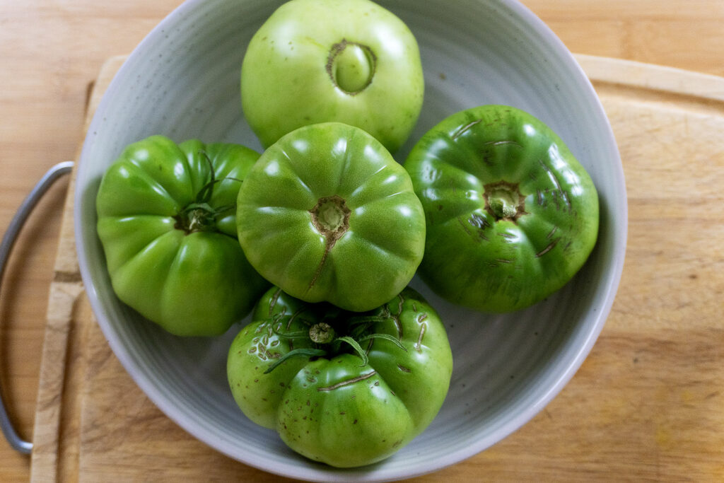A white bowl filled with five large, unripe green tomatoes sits on a wooden surface next to a cutting board.