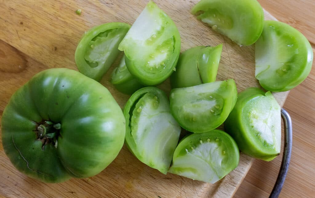 A whole green tomato and several wedges of sliced green tomato are on a wooden cutting board.