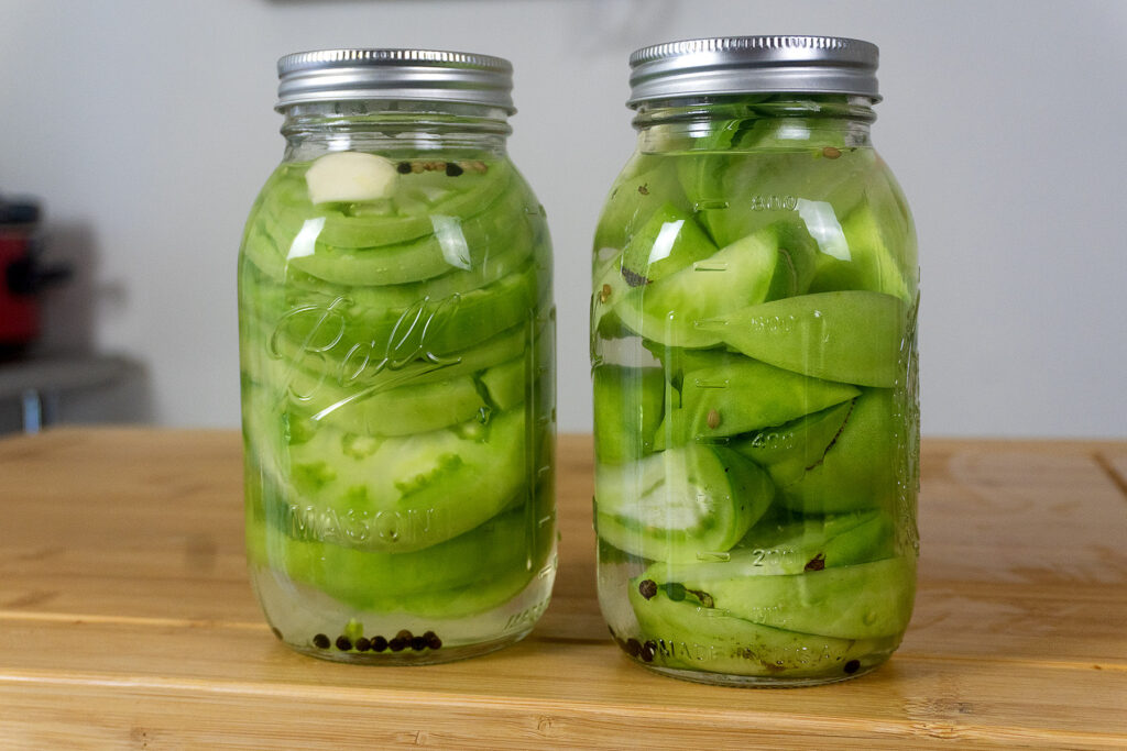 Two glass jars filled with sliced green tomatoes, garlic cloves, peppercorns, and pickling liquid sit on a wooden surface. Both jars are sealed with metal lids.