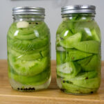 Two glass jars filled with sliced green tomatoes, garlic cloves, peppercorns, and pickling liquid sit on a wooden surface. Both jars are sealed with metal lids.
