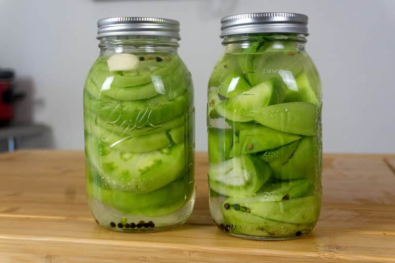 Two glass jars filled with sliced green tomatoes, garlic cloves, peppercorns, and pickling liquid sit on a wooden surface. Both jars are sealed with metal lids.