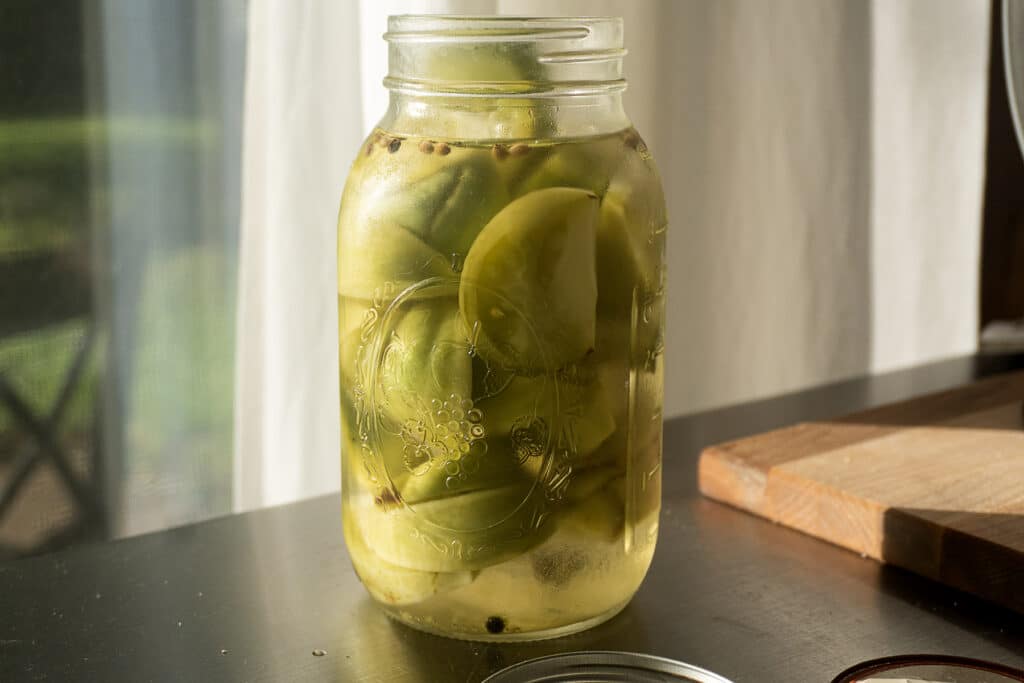 A glass jar filled with green apple slices in brine sits on a dark table near a window, with sunlight shining through. A wooden cutting board is partially visible in the background.