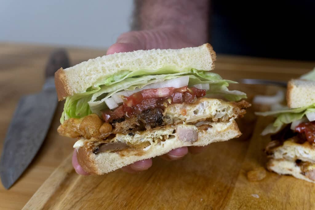 A hand holds a sandwich with lettuce, tomato, onion, fried egg, and various meats between two slices of white bread. A knife lies on a wooden cutting board in the background.