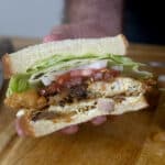 A hand holds a sandwich with lettuce, tomato, onion, fried egg, and various meats between two slices of white bread. A knife lies on a wooden cutting board in the background.