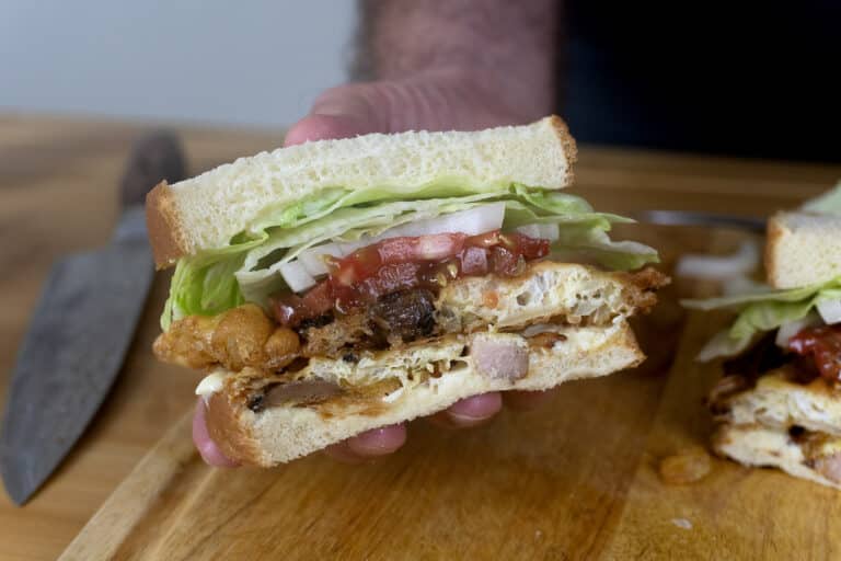 A hand holds a sandwich with lettuce, tomato, onion, fried egg, and various meats between two slices of white bread. A knife lies on a wooden cutting board in the background.