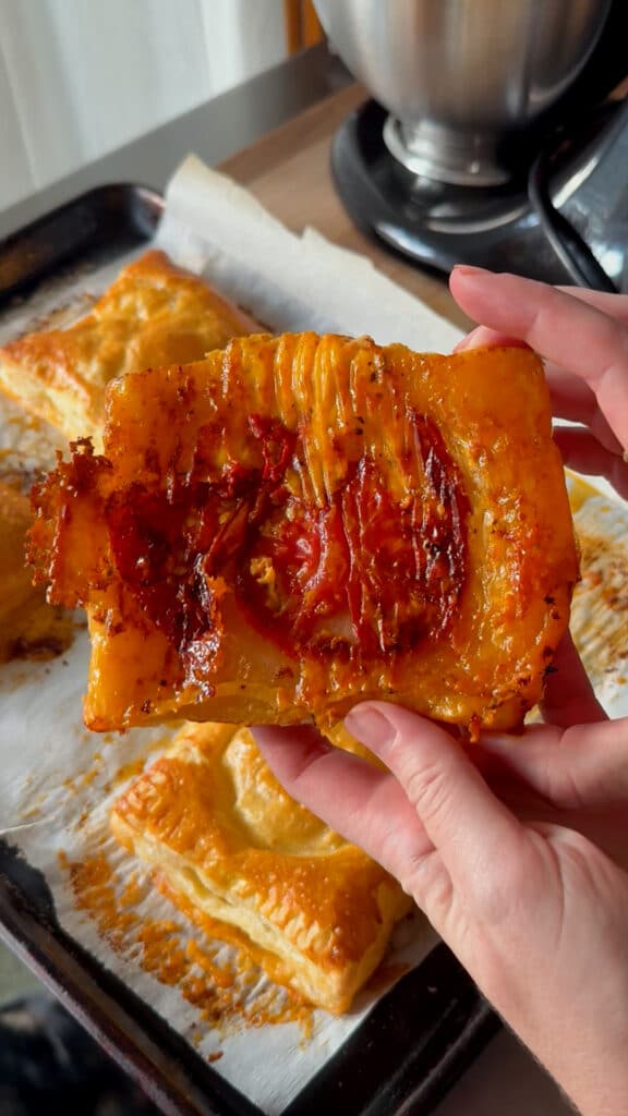 A hand holds a rectangular piece of puff pastry topped with roasted tomato slices and herbs, with more pastries on a parchment-lined baking tray in the background.