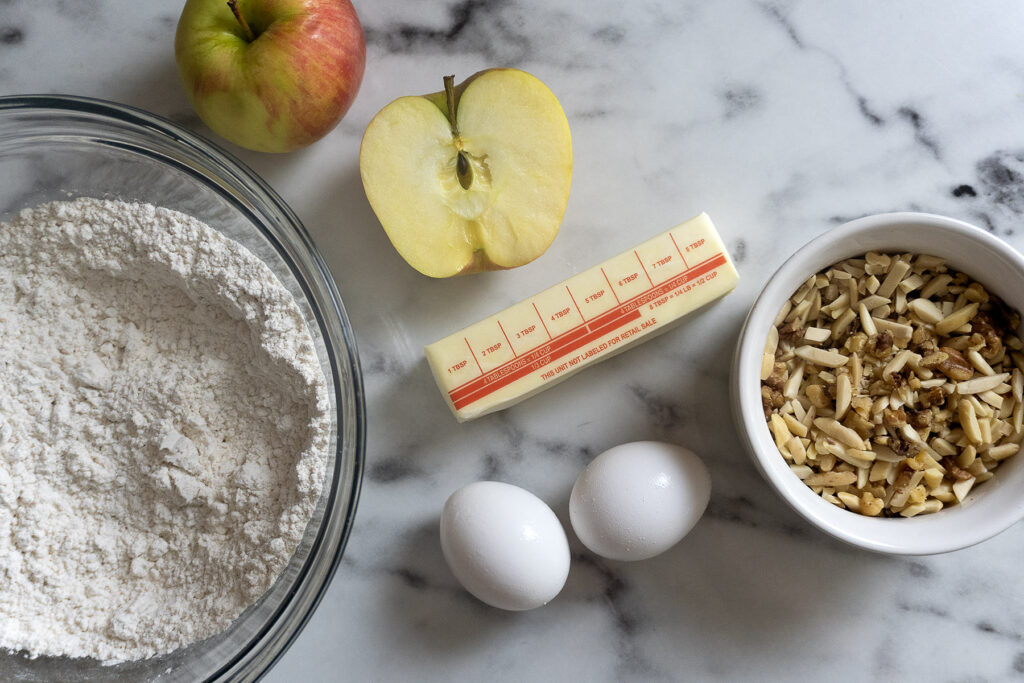 Bowl of flour, whole apple, apple half, stick of butter, two eggs, and bowl of chopped nuts on a marble surface.