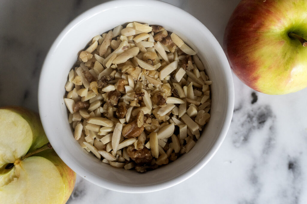 A white bowl filled with chopped walnuts and slivered almonds sits on a marble surface next to a whole apple and a sliced apple half.
