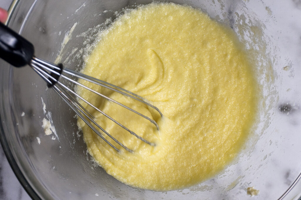 A whisk sits in a glass bowl filled with a thick, yellow batter being mixed.