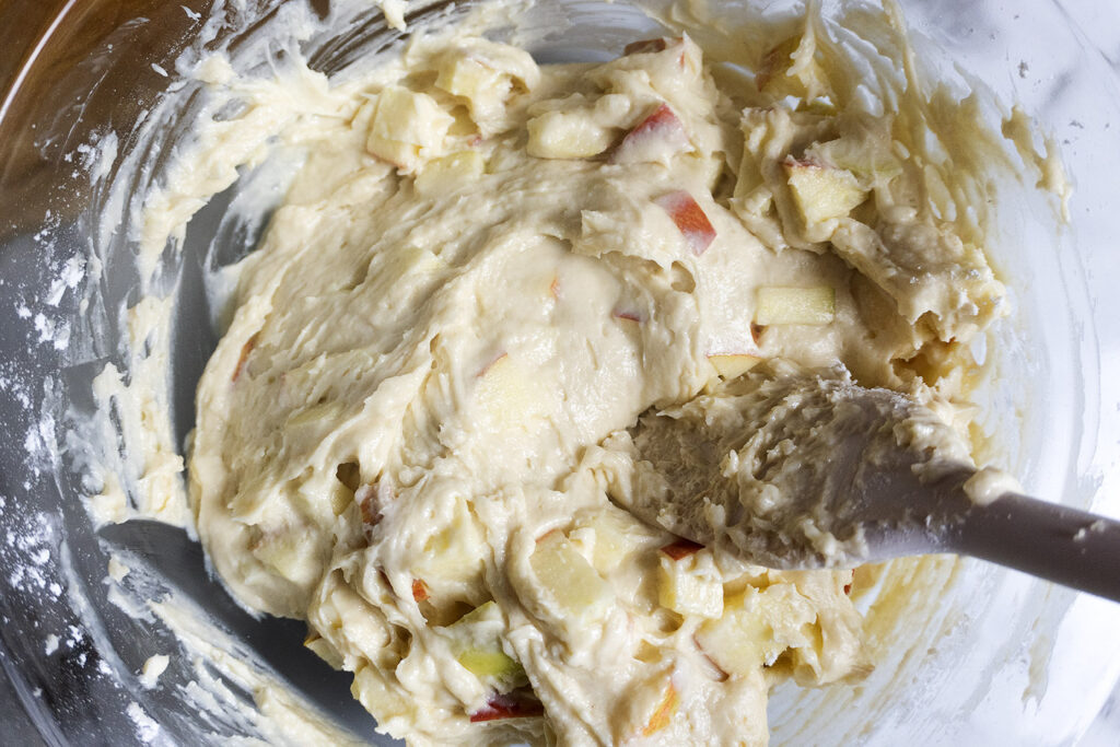 A glass bowl filled with thick cake batter containing apple chunks, being stirred with a spatula.