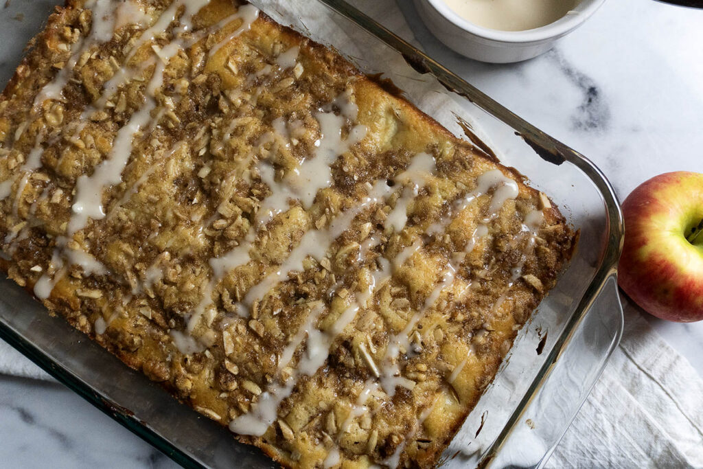 A glass baking dish holds apple crumble cake with icing drizzled on top. An apple and a bowl of icing sit on a marble countertop nearby, with a white towel partially visible underneath the dish.