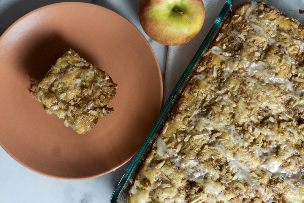 A slice of apple crumb cake on a peach plate, next to a whole apple and a glass baking dish filled with the remaining cake, topped with icing and crumbs.
