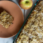 A slice of apple crumb cake on a peach plate, next to a whole apple and a glass baking dish filled with the remaining cake, topped with icing and crumbs.