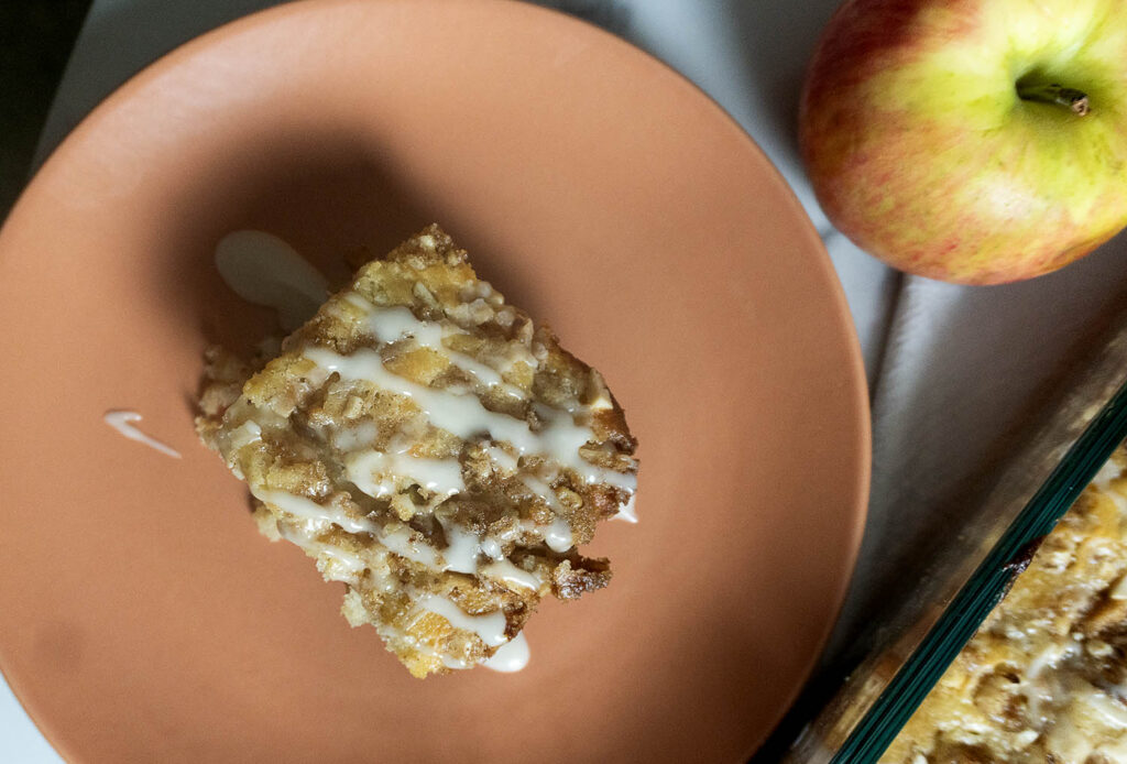 A slice of apple crumb cake with icing drizzle on a peach-colored plate, next to a whole apple and part of a baking dish.