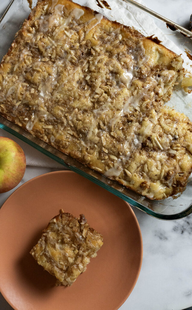 A glass baking dish with a large apple cake partially cut, topped with icing and crumbs. A square slice is on a pink plate nearby, with a fresh apple beside it on a marble surface.