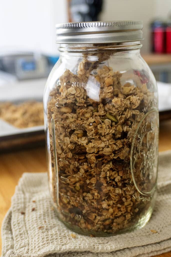 A mason jar filled with homemade granola sits on a beige cloth, with a blurred baking tray and kitchen items in the background.