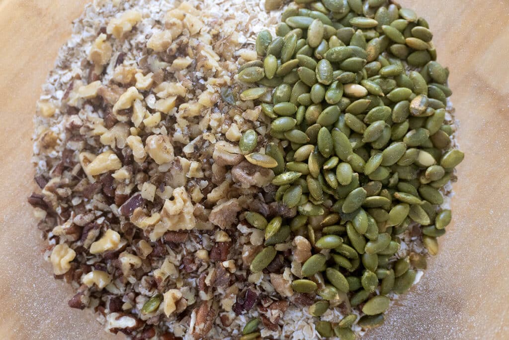 Chopped walnuts and pumpkin seeds sit side by side on a bed of oats in a wooden bowl, ready to be mixed for a recipe.