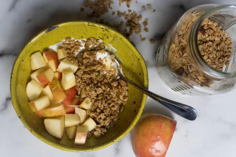 A yellow bowl filled with yogurt, chopped apples, and granola, with a spoon inside. A jar of granola, scattered granola pieces, and an apple are beside the bowl on a white surface.