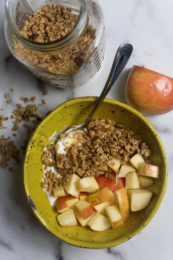 A yellow bowl with yogurt, granola, and chopped apples, next to a glass jar of granola and a red apple on a white marble surface. A spoon is in the bowl and some granola is scattered nearby.