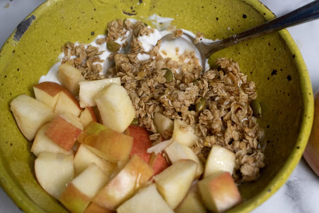 A green bowl contains chopped apples, granola, and yogurt, with a metal spoon resting inside.