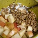 A green bowl contains chopped apples, granola, and yogurt, with a metal spoon resting inside.