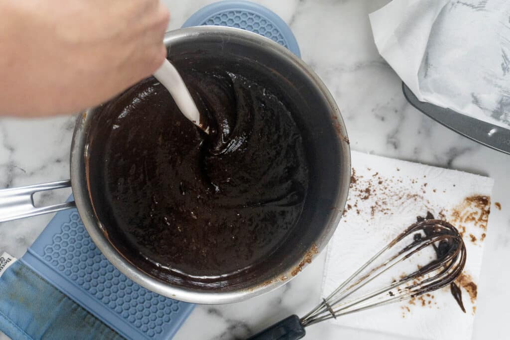 A hand uses a white spatula to stir thick, dark chocolate batter in a metal saucepan sitting on a blue silicone trivet, with a wire whisk on a paper towel nearby.