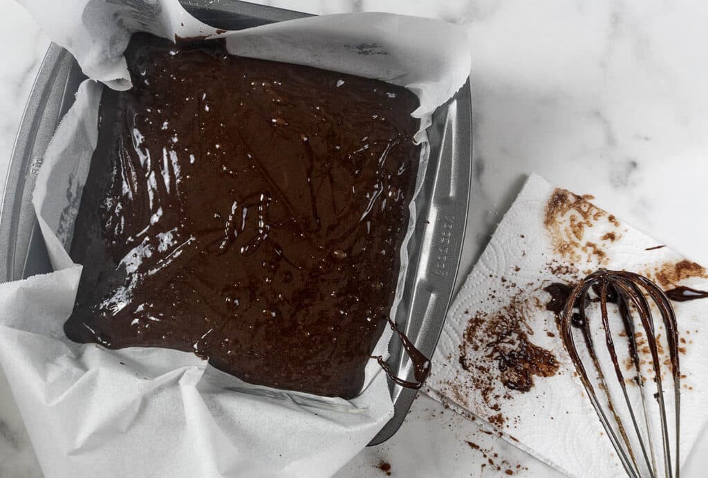 A square baking pan filled with chocolate brownie batter, next to a whisk resting on a stained paper towel.