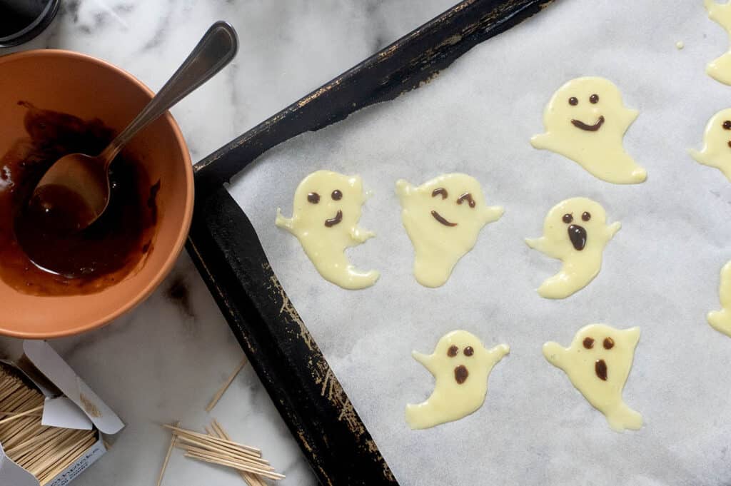 Ghost-shaped pancake batter on a parchment-lined baking sheet, with melted chocolate and toothpicks nearby for drawing faces.