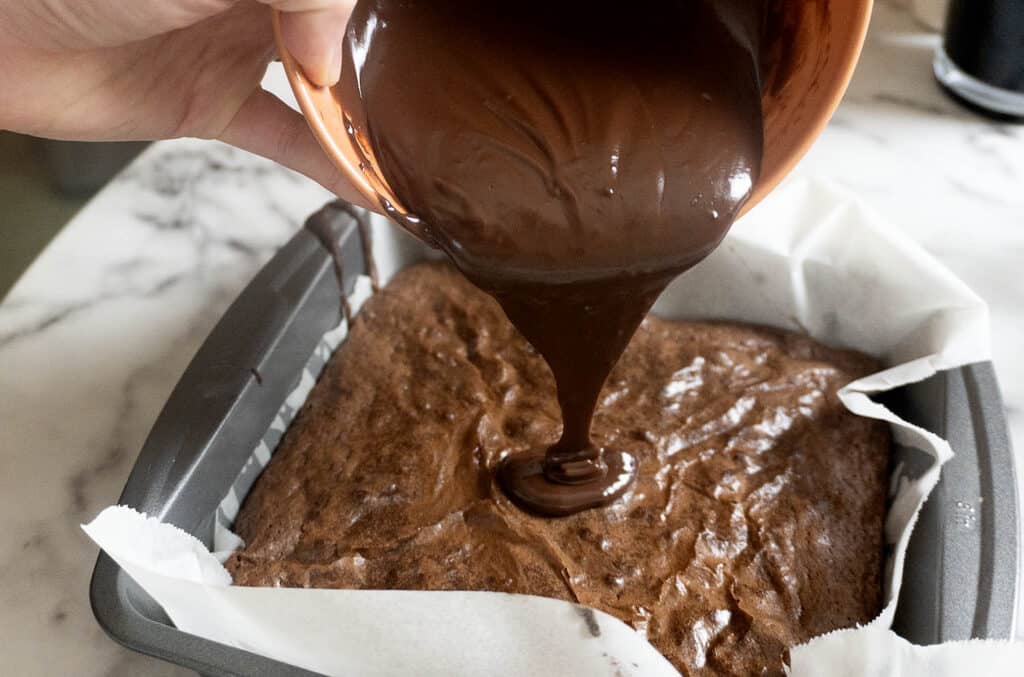Chocolate ganache being poured from a bowl onto a baked brownie in a square pan.