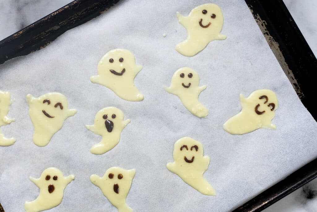 Nine white chocolate ghost shapes with chocolate-drawn faces on a parchment-lined baking sheet.