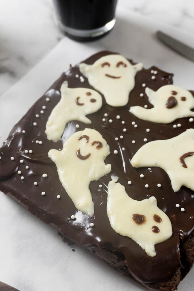 A square chocolate brownie frosted with chocolate ganache and decorated with white chocolate ghosts and white sprinkles.