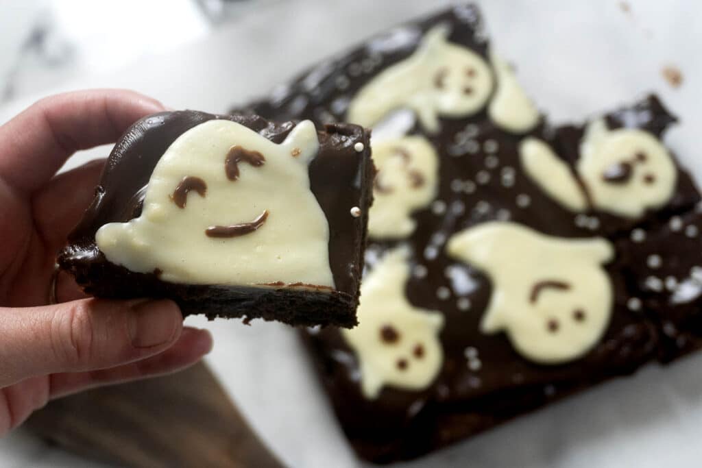 A hand holds a chocolate brownie decorated with a white chocolate ghost, with more ghost-topped brownies on a board in the background.
