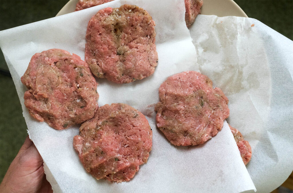 Five raw, seasoned meat patties resting on parchment paper on a plate.