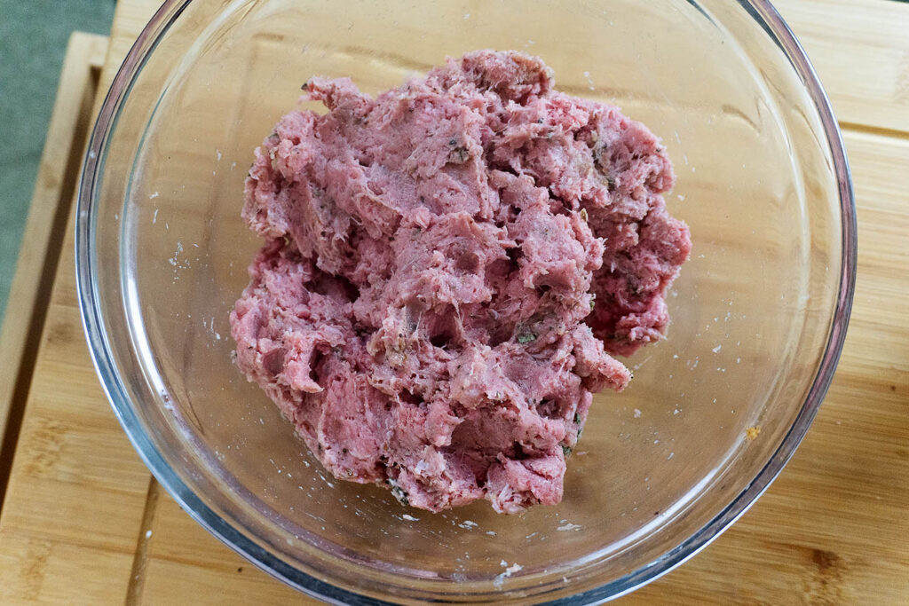 A glass bowl filled with raw ground meat mixed with herbs on a wooden cutting board.